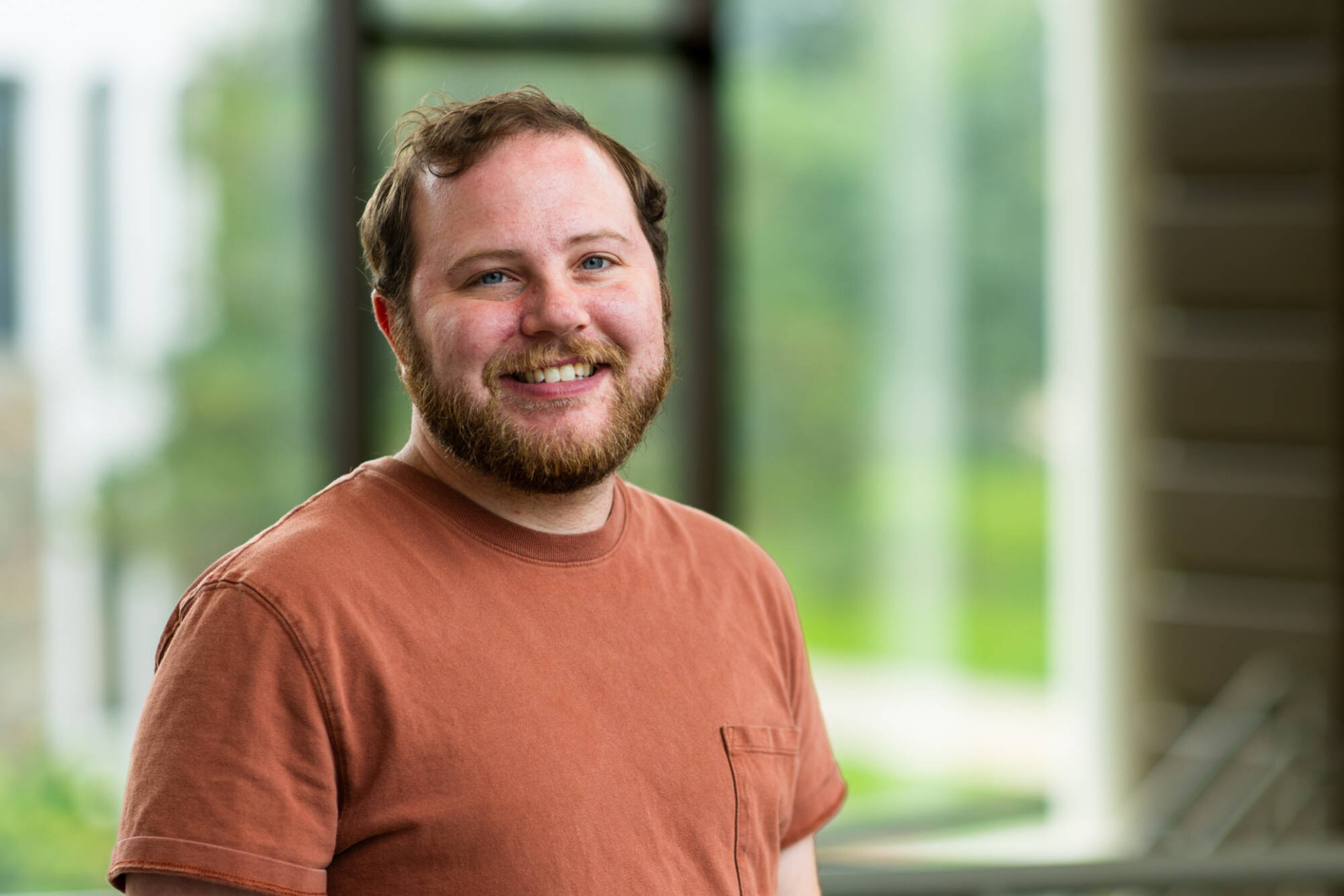 A smiling man with dark hair and a beard, wearing an orange shirt, standing in front of a blurred green background.